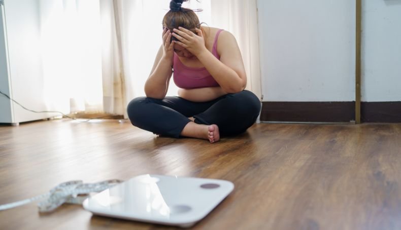 Mujer con Sobrepeso Sentada en el Piso Cerca de una Balanza con Manos en la Cabeza en Señal de Frustración